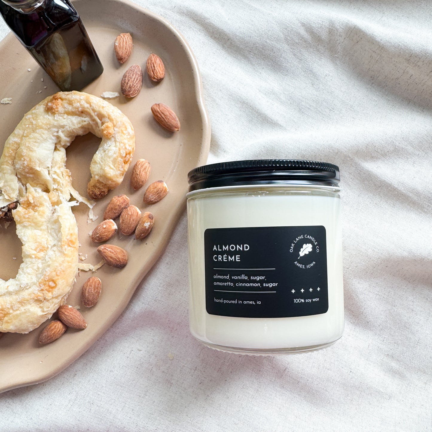 Candle labeled 'Almond Cèreme' next to almonds and a bagel on a white surface