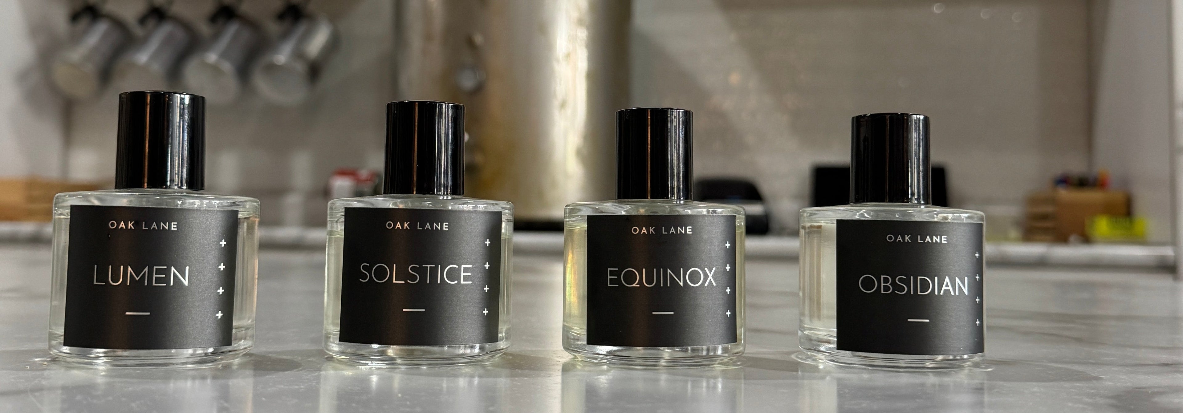 A close-up of several small perfume or fragrance bottles with black labels and black caps, mounted on a clear display stand inside a white box. The box has the text 'SOMETHING SMELLS amazing IN HERE' printed on it.